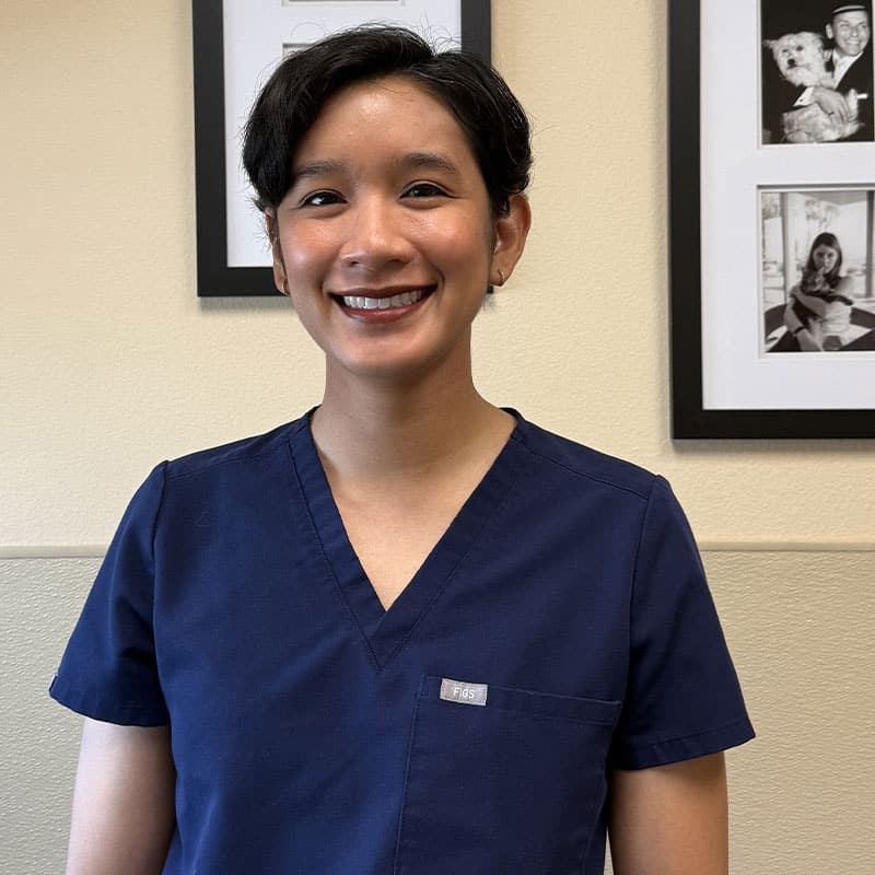 A person with short dark hair, wearing navy blue medical scrubs, smiles while standing indoors. Black-and-white photos in black frames are visible on the beige wall behind them.