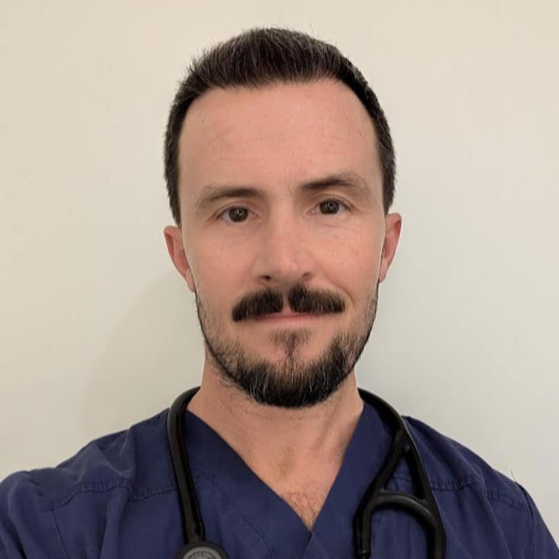 A man with short dark hair and a trimmed beard, wearing navy blue scrubs and a stethoscope around his neck, stands in front of a plain light-colored wall.