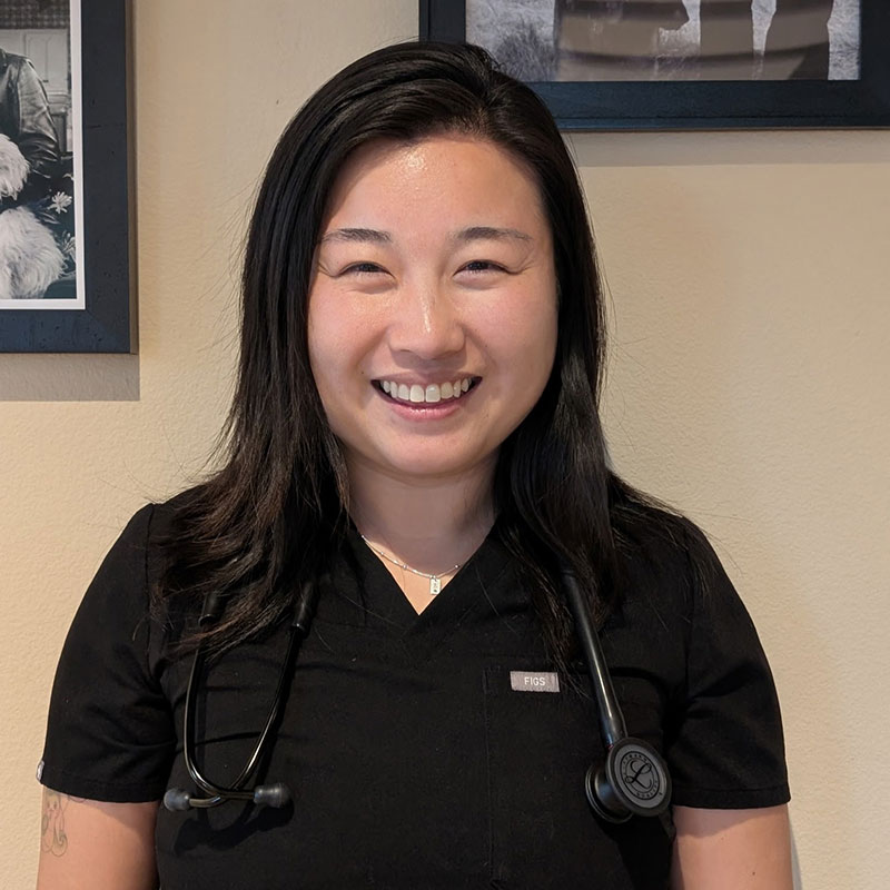 A woman with long dark hair, wearing black scrubs and a stethoscope around her neck, smiles at the camera. She is standing indoors in front of a beige wall with framed photos.